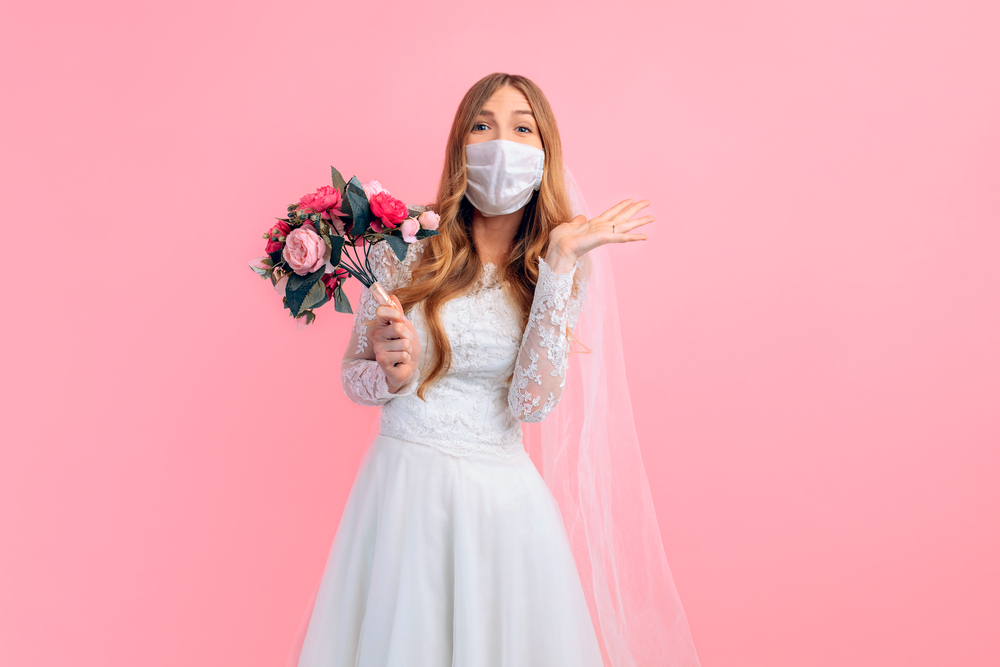 Bride wearing medical face mask and holding a bouquet of flowers in front of a pink background.